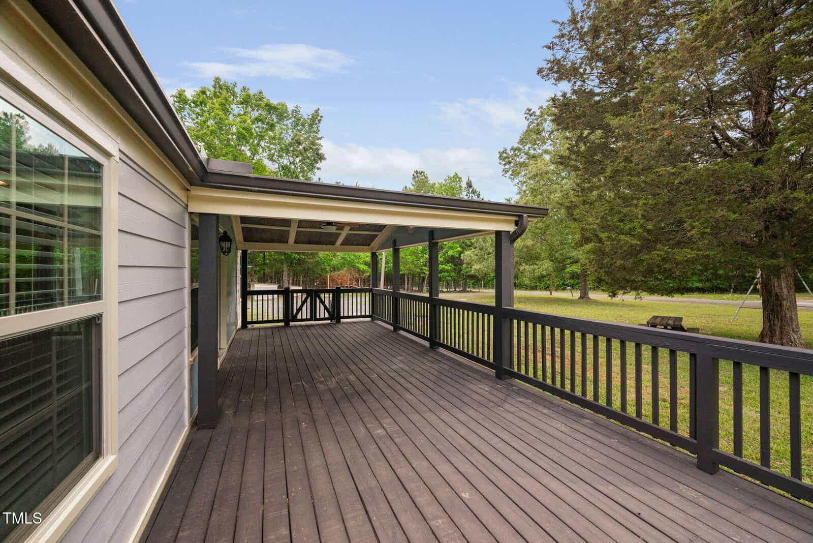 A spacious covered wooden deck with black railings attached to a house, overlooking a grassy yard with trees under a partly cloudy sky.
