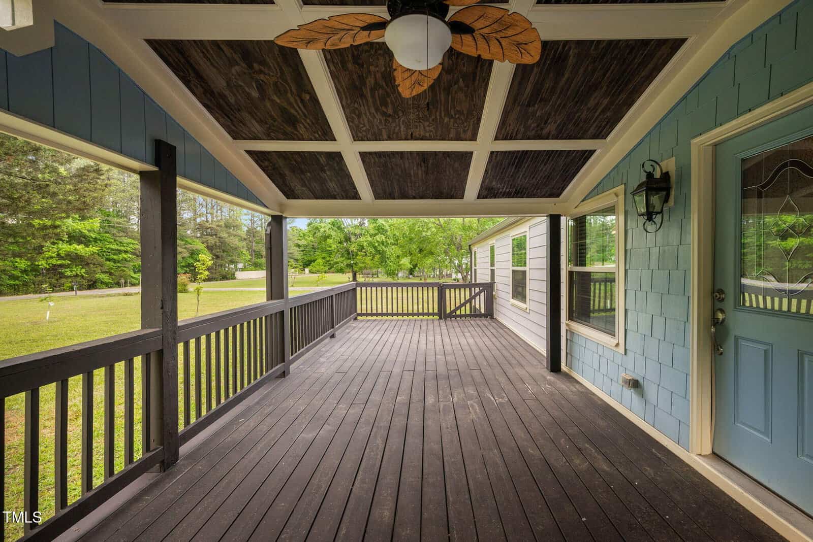 A covered front porch with dark wooden flooring and railings, a ceiling fan with leaf-shaped blades, blue siding, a glass-paneled door, and a lantern-style light fixture. Trees and grass are visible outside.
