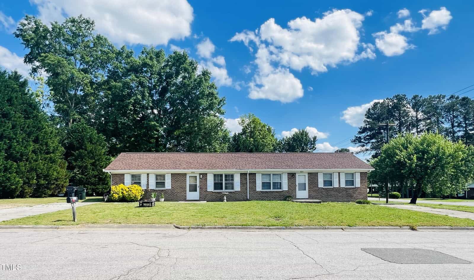 A one-story brick duplex with white trim sits on a grassy lot next to a street. The building features four white doors and several windows. Trees and a clear blue sky with scattered clouds are in the background.