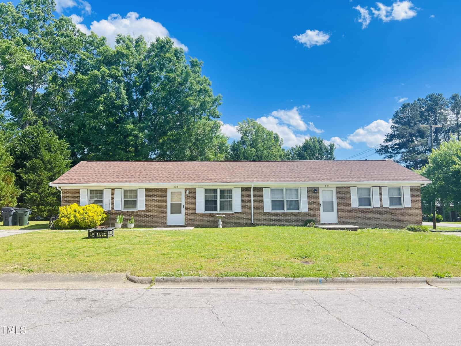 A charming one-story brick duplex at 805-807 Sunset Avenue, Oxford, featuring a red shingle roof, white trim, and four front doors. It sits on a grassy lawn with shrubs and trees under a bright blue sky.