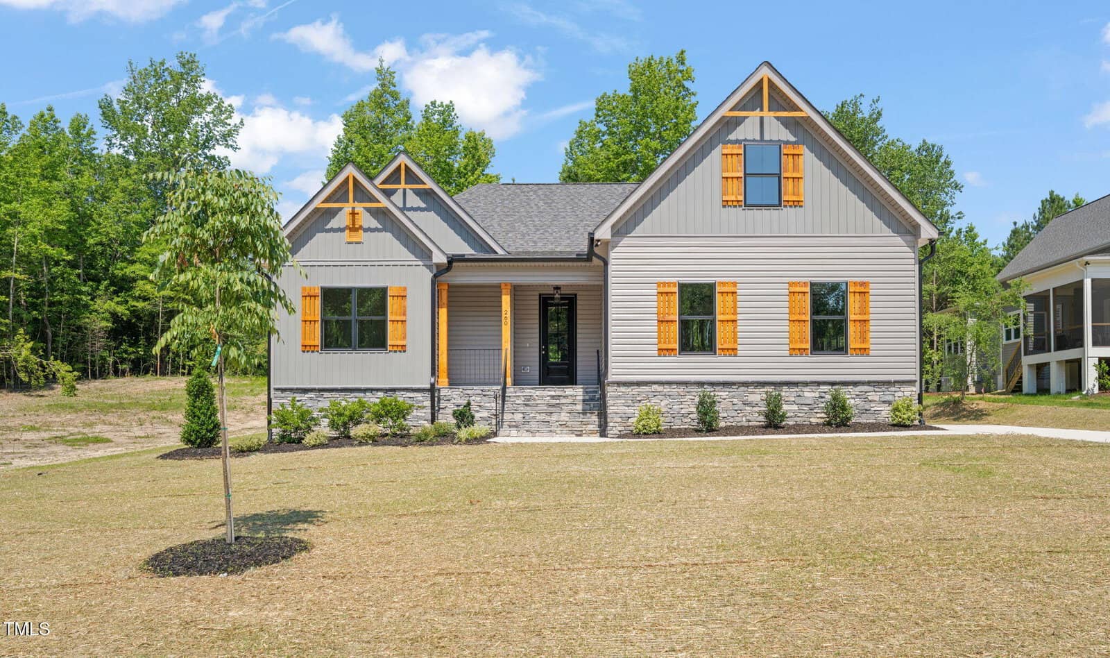 A modern gray house at 260 Pilot Ridge Road in Zebulon features yellow wooden shutters and stone accents, sitting on a newly landscaped lawn with young trees, all surrounded by greenery under a blue sky with scattered clouds.