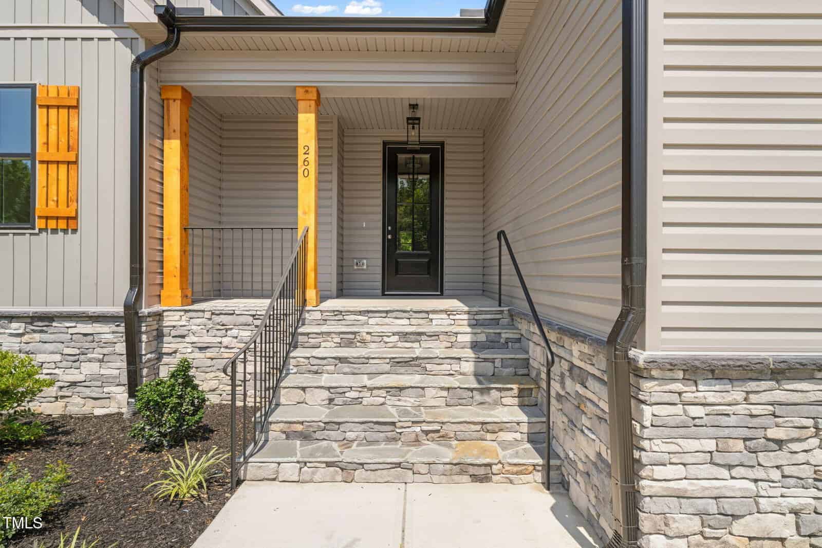 A front porch with stone steps, black metal railings, beige siding, stone veneer, wooden posts, and a black door with glass panel. A small garden with shrubs lines the walkway.