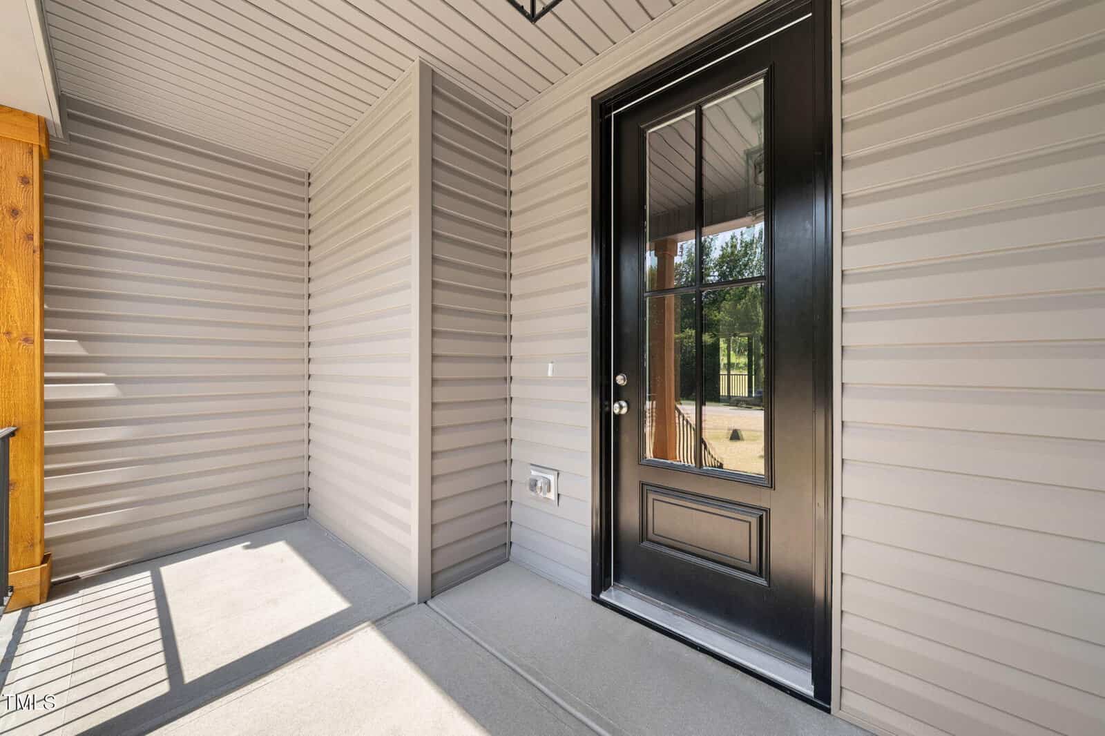 Covered front porch with light gray vertical siding, a black-framed glass panel door, and concrete flooring. Sunlight casts shadows through the open area onto the porch floor.