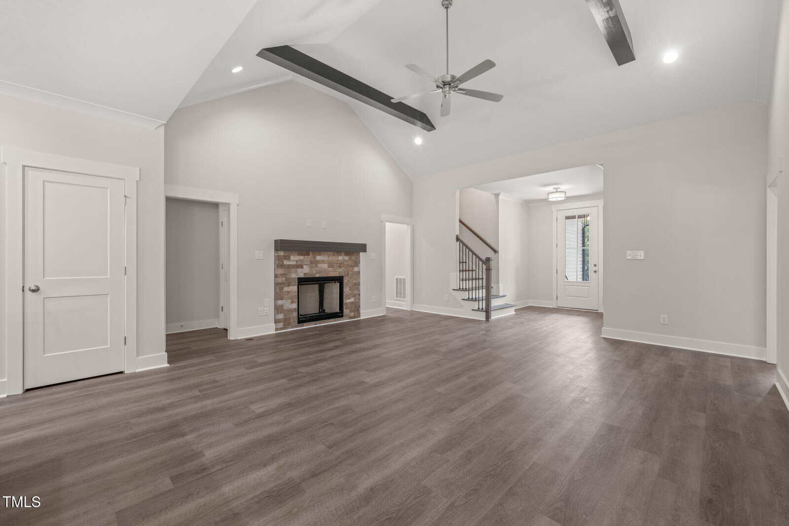 Spacious living room with vaulted ceiling, exposed beams, ceiling fan, light gray walls, wood-style flooring, a fireplace with a brick surround, and entryway opening to a staircase and front door.