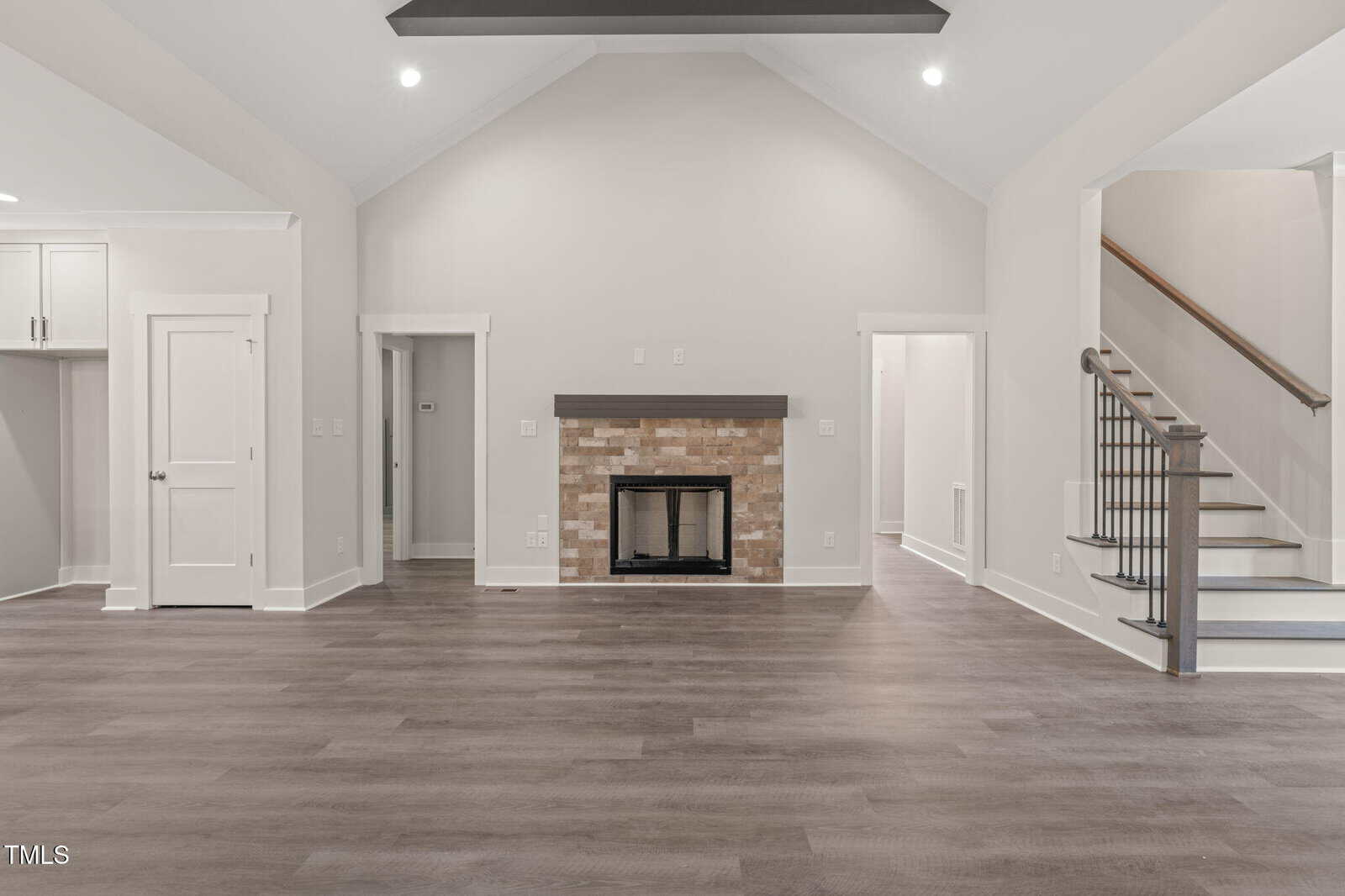 Spacious living room with light gray walls, vaulted ceiling, wood-style flooring, a fireplace with a brick surround, and staircase with dark railing leading upstairs. White trim and recessed lighting add a modern touch.