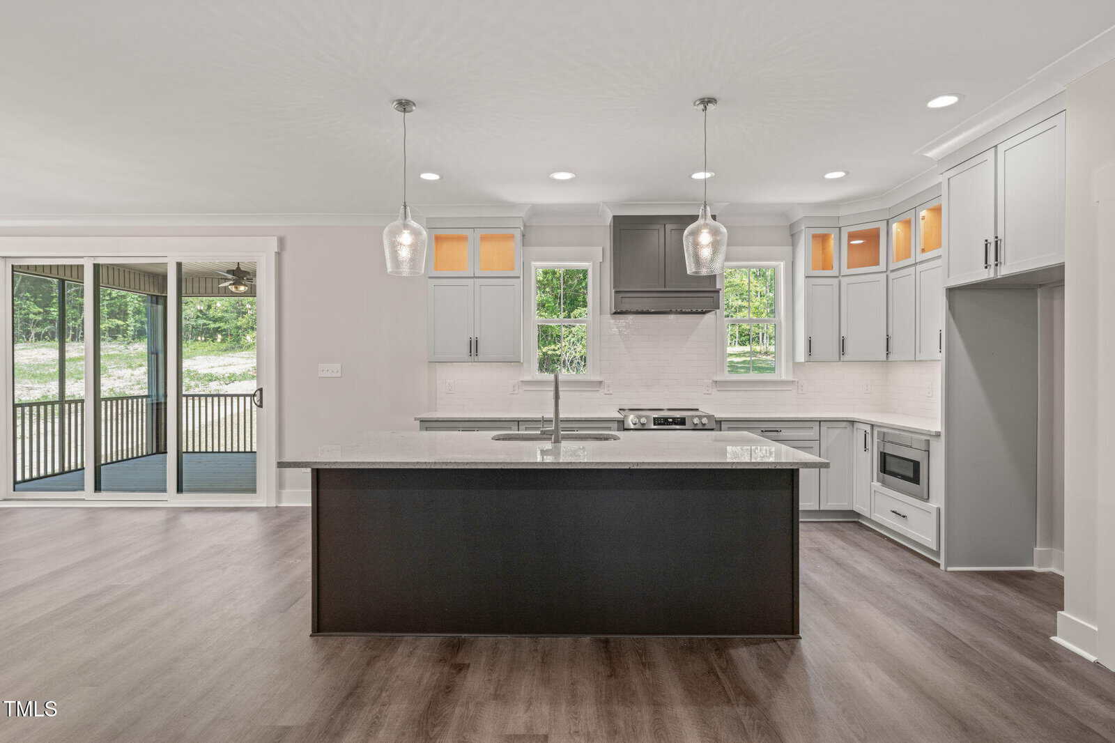 Modern kitchen with gray cabinets, stainless steel appliances, a large island with sink, pendant lights, and light countertops. Sliding doors open to a covered patio with a wooded view outside.