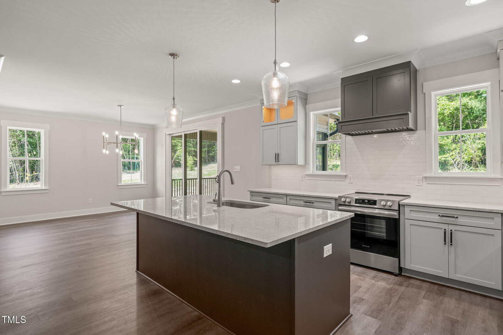 Modern kitchen with gray cabinets, stainless steel appliances, an island with a sink, pendant lights, and large windows providing natural light. Open floor plan extends to a dining area with wood flooring and outdoor views.