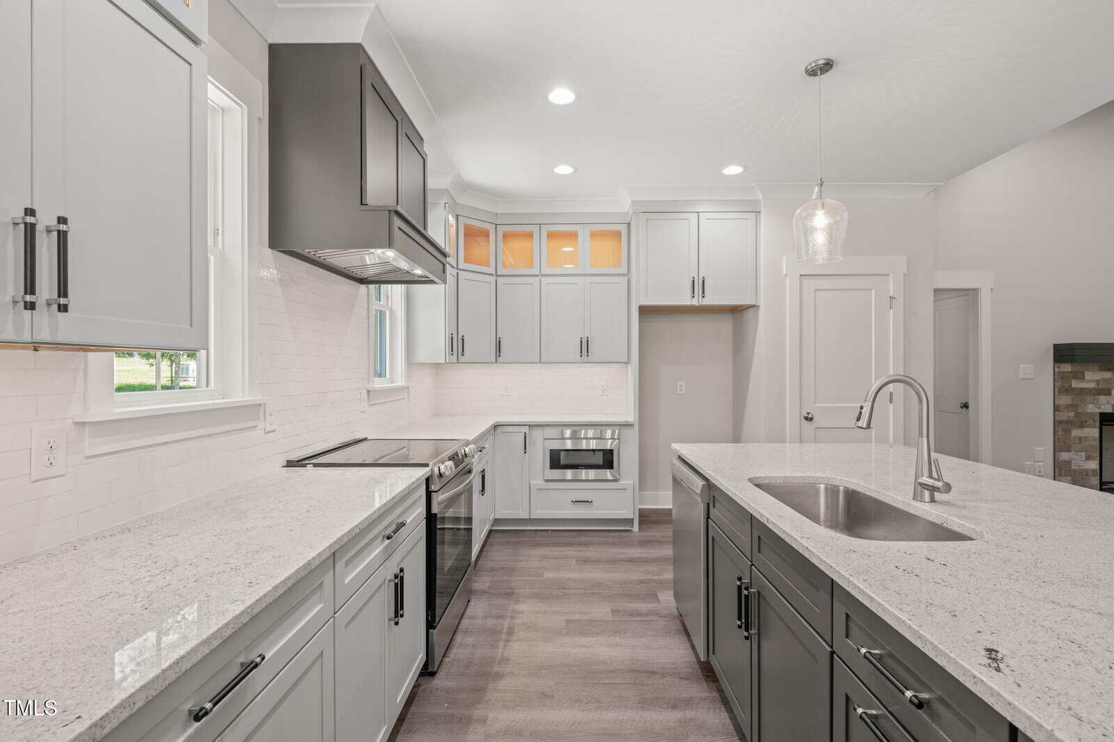 Modern kitchen with white and gray cabinets, stainless steel appliances, a large island with a sink, pendant lighting, and light gray countertops. The space is bright with natural and overhead light.