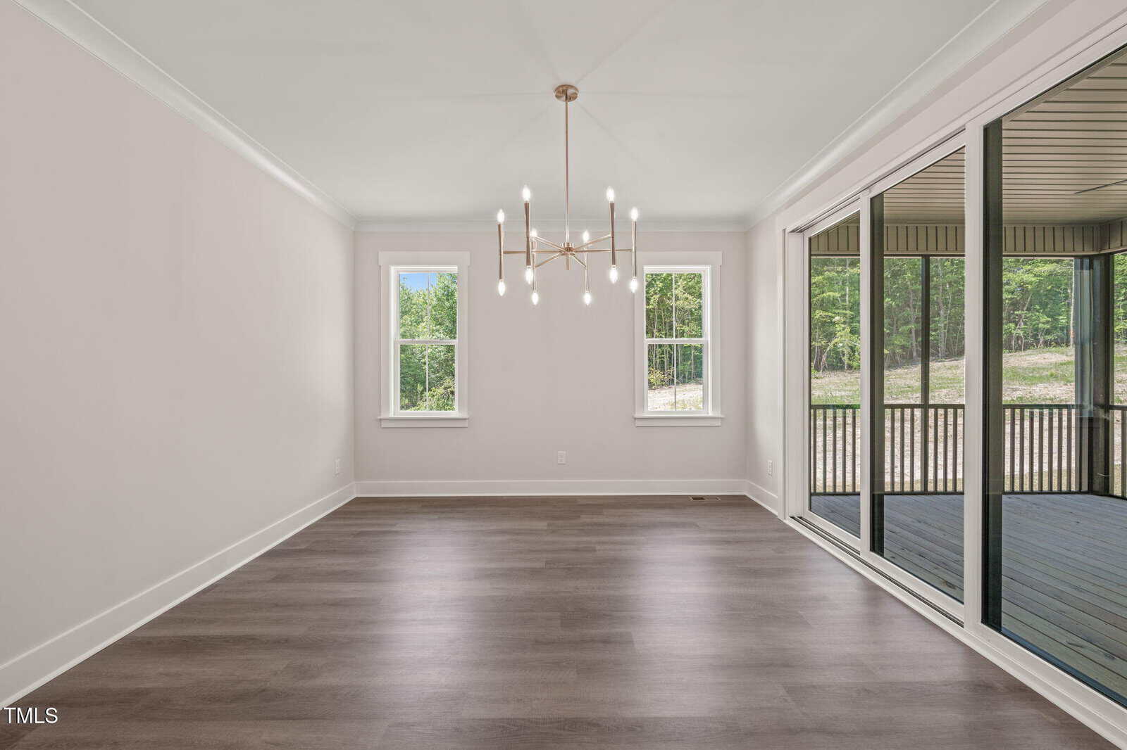 Bright, empty room with light walls, wide plank wood flooring, modern chandelier, two windows, and large sliding glass doors opening to a screened porch with a wooded view outside.