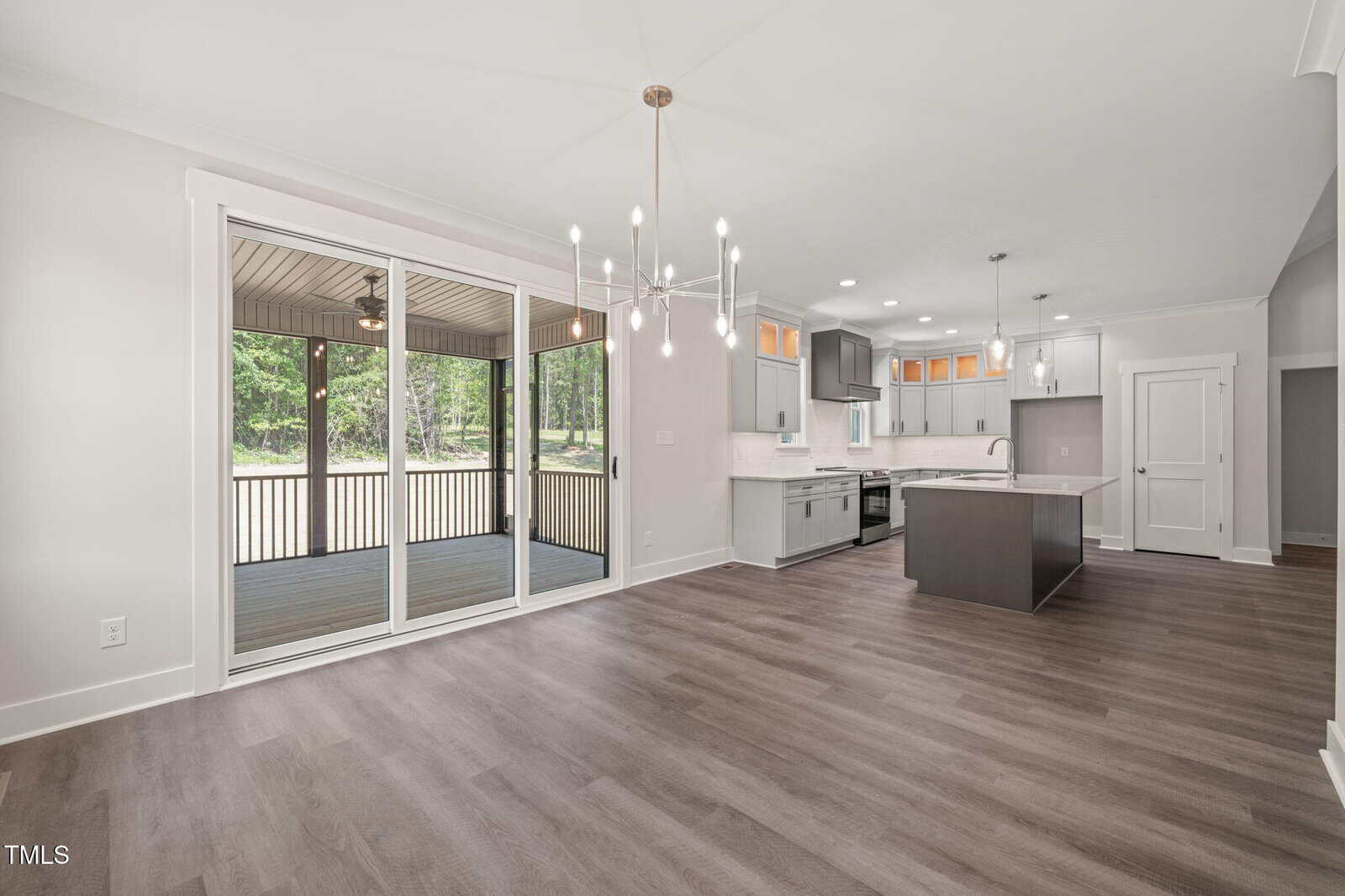 Modern open-concept kitchen and dining area with wood flooring, white cabinets, pendant lights, and large sliding glass doors leading to a screened porch with a wooded view.