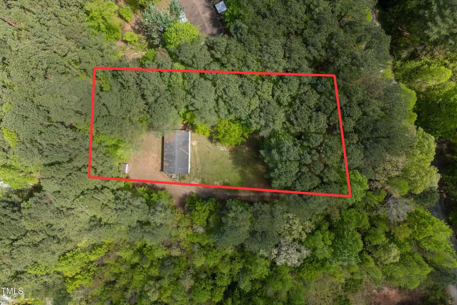 Aerial view of a house surrounded by dense trees, with a red outline marking the property boundary. The house has a brown roof and is situated near the center of the outlined area.