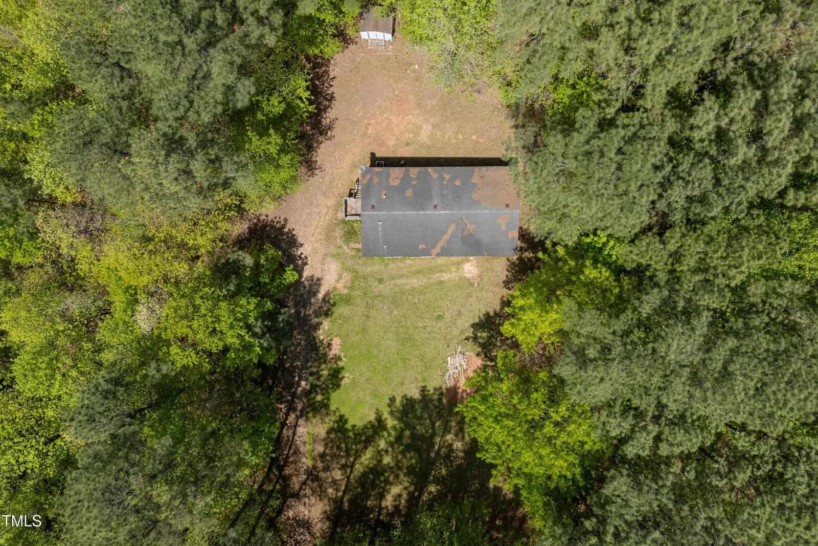 Aerial view of a rectangular building with a dark roof surrounded by dense trees and a patch of grass, with a small shed visible near the top edge of the clearing.