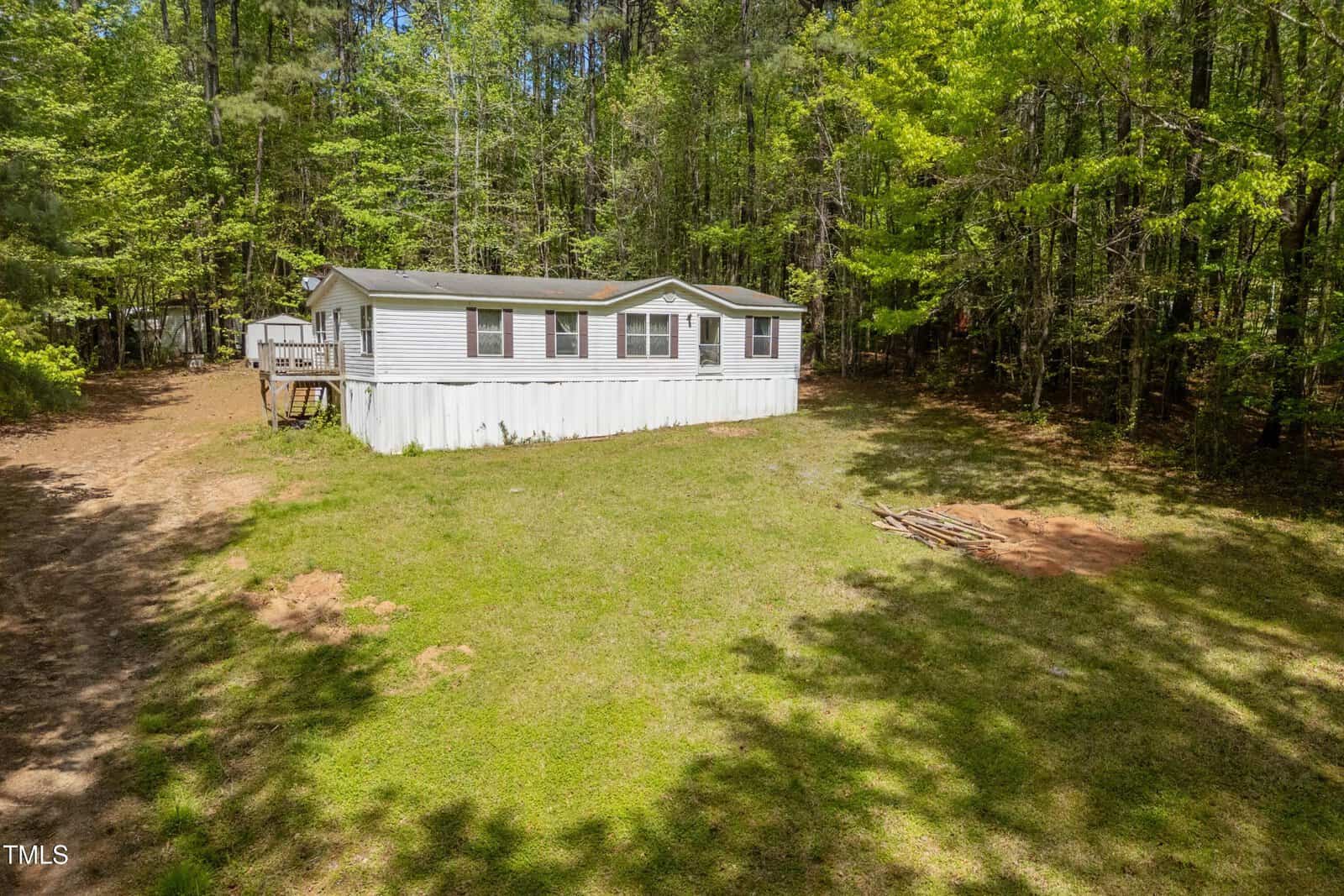 A single-story white manufactured home with a small deck sits on a grassy clearing at 3143 Tree Top Drive, Wake Forest, surrounded by dense trees. A small pile of branches rests on the lawn, with a shed partially visible in the background.