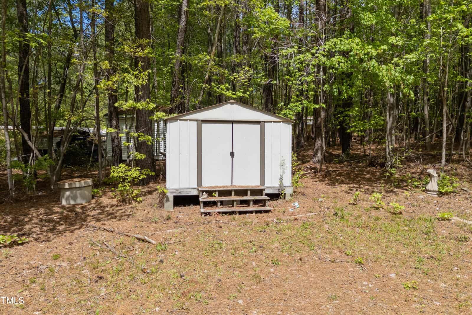 A white storage shed with a metal roof and double doors stands on a patch of bare ground, surrounded by trees and greenery. A small wooden step leads to the entrance.