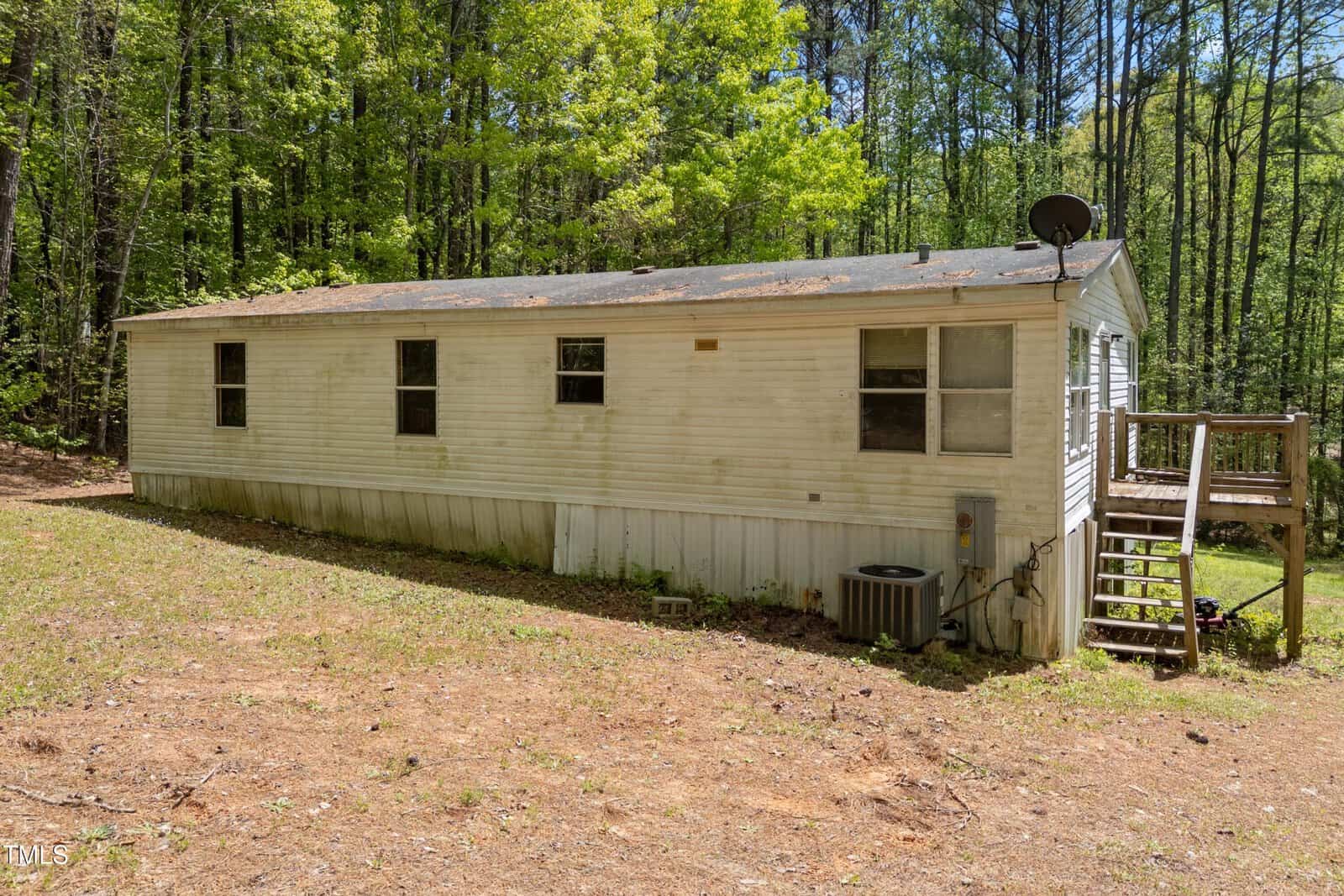 A white single-wide mobile home with a shingled roof and several windows is set on a grassy, wooded lot. There is a small wooden stairway at the entrance and an air conditioning unit on the side.