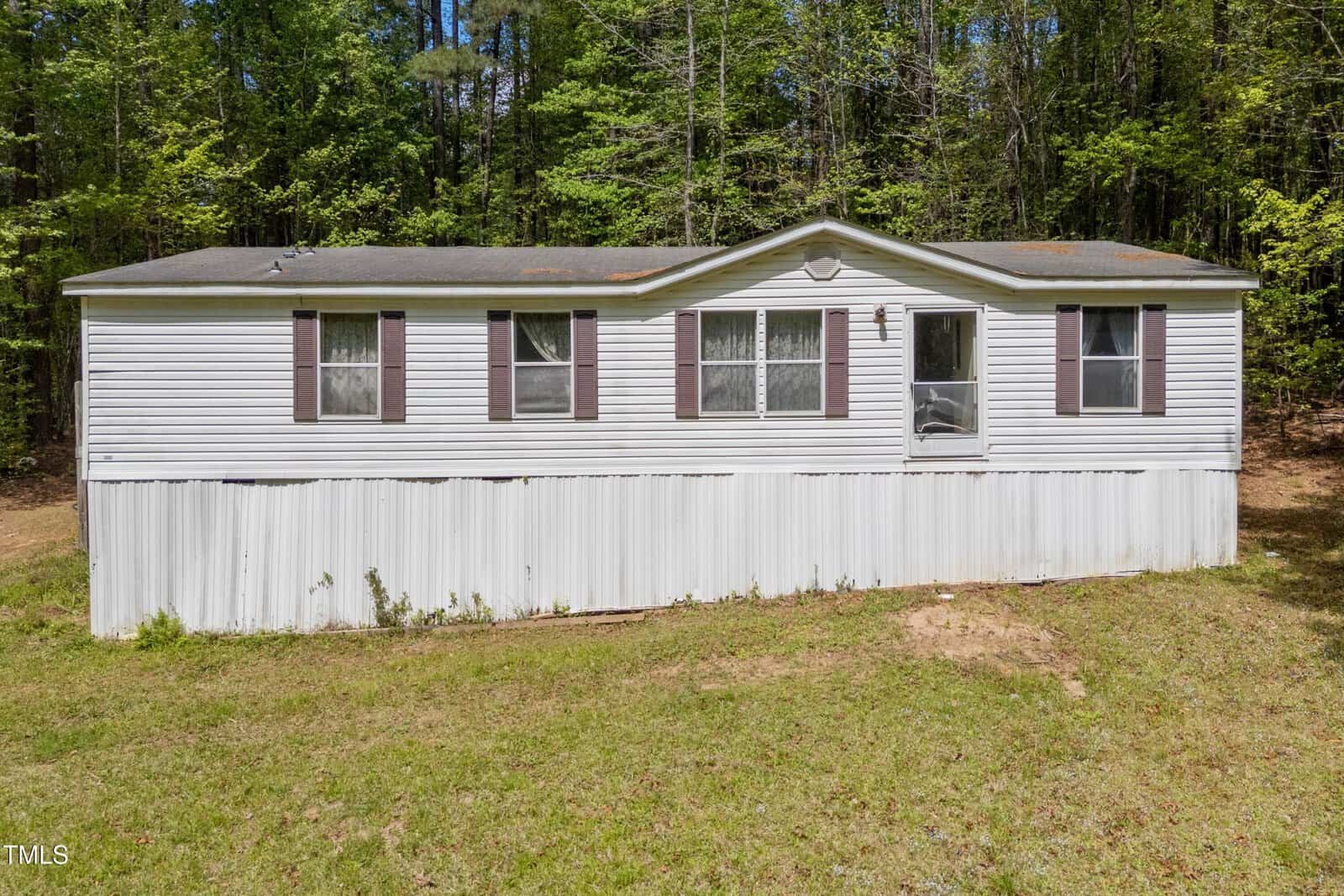 A single-story white mobile home with a dark shingle roof, multiple windows with brown shutters, and a single front door, set on a grassy lot with trees in the background.