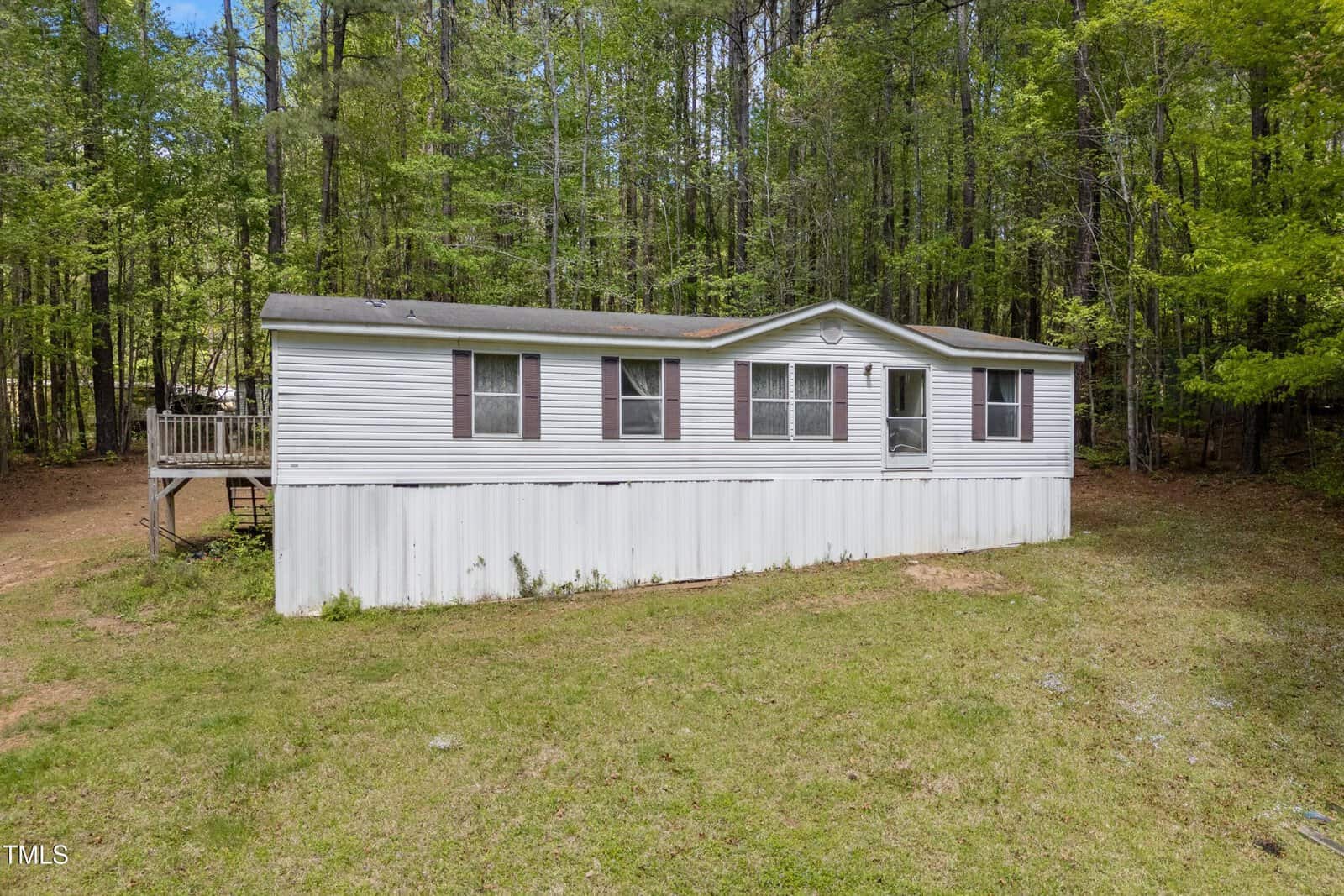 A white single-wide mobile home with several windows and a small porch on the left side, set on a grassy lot bordered by tall trees in the background.