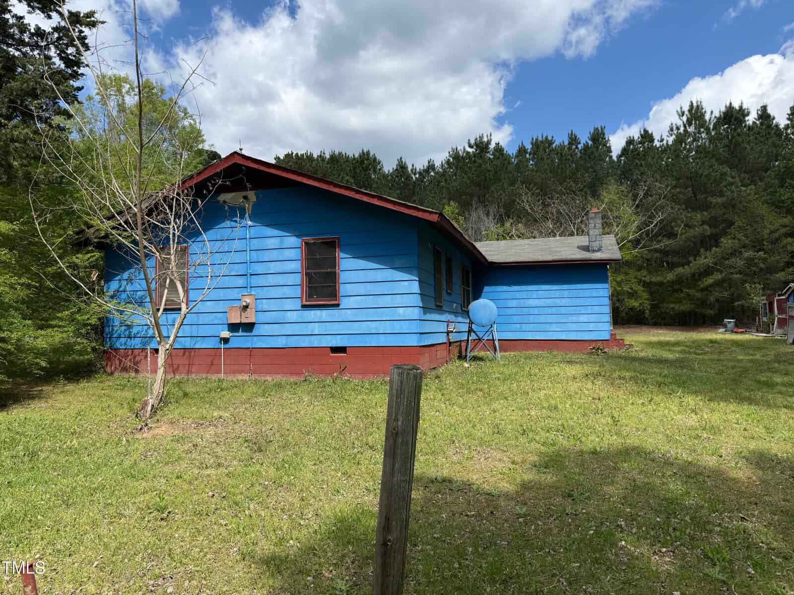 A small, single-story blue house with a red foundation sits on a grassy lawn bordered by trees. A satellite dish is attached to the side of the house, and the sky is partly cloudy.