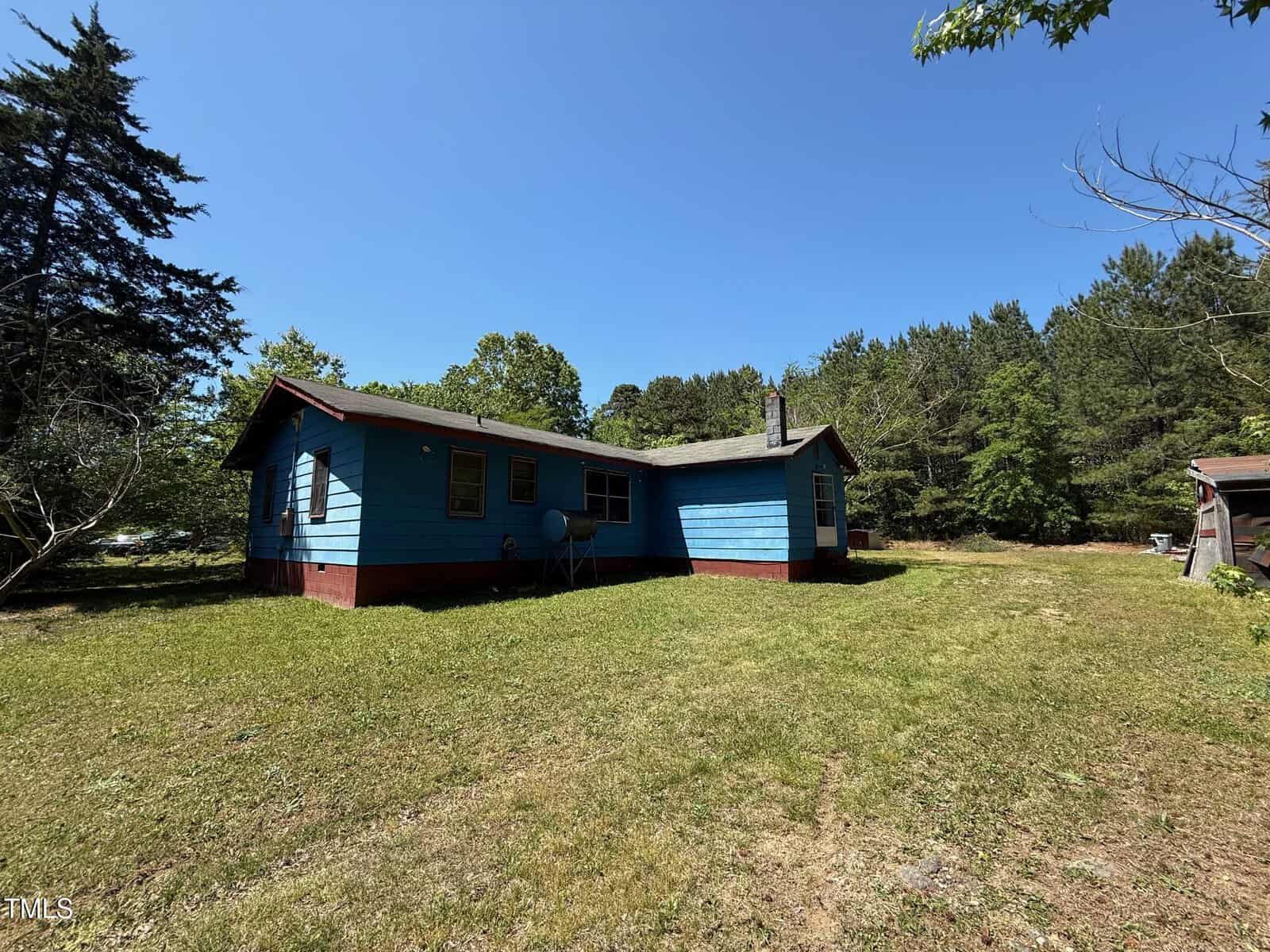 A small, single-story blue house with a red foundation sits on a grassy lawn surrounded by trees under a clear blue sky. A satellite dish is attached to the house, and a shed is partially visible on the right.