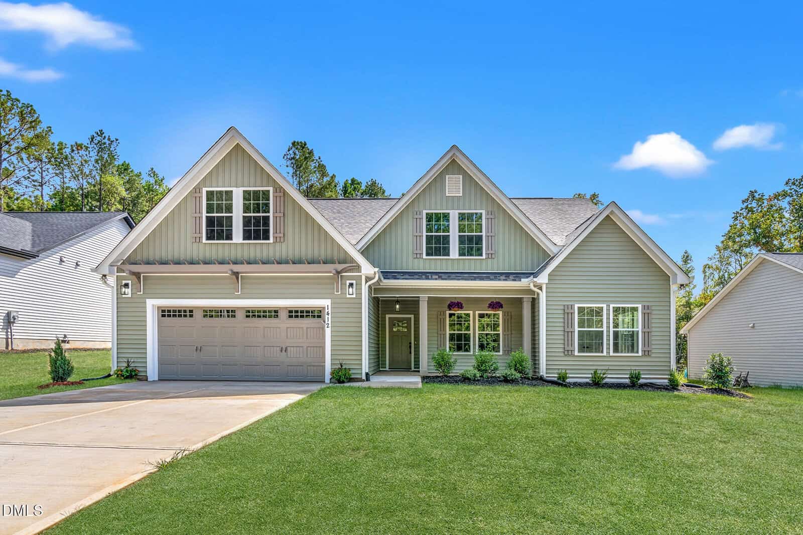 A modern light green house at 1412 Sagamore Drive, Louisburg, featuring a two-car garage, gabled roof, front porch, and well-kept lawn. A concrete driveway leads to the garage; clear blue sky and trees are in the background.