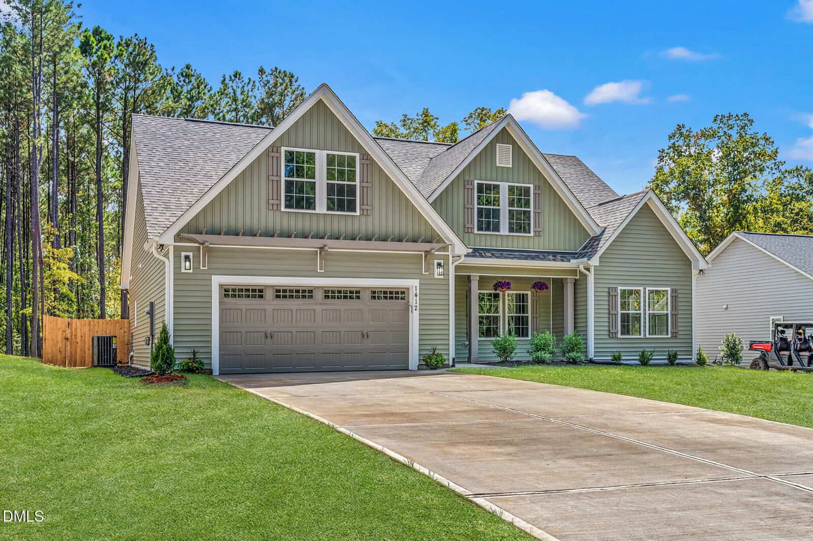 A two-story suburban house with gray siding, a double garage, a large driveway, and a well-kept lawn. There are trees in the background and a clear blue sky overhead.