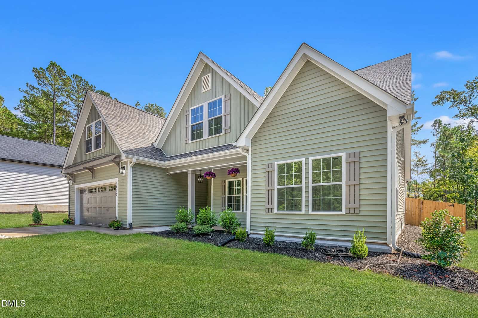 A light green two-story suburban house with a gabled roof, attached two-car garage, shuttered windows, hanging flower baskets, and a well-maintained lawn with shrubs and mulch. The sky is clear and blue.