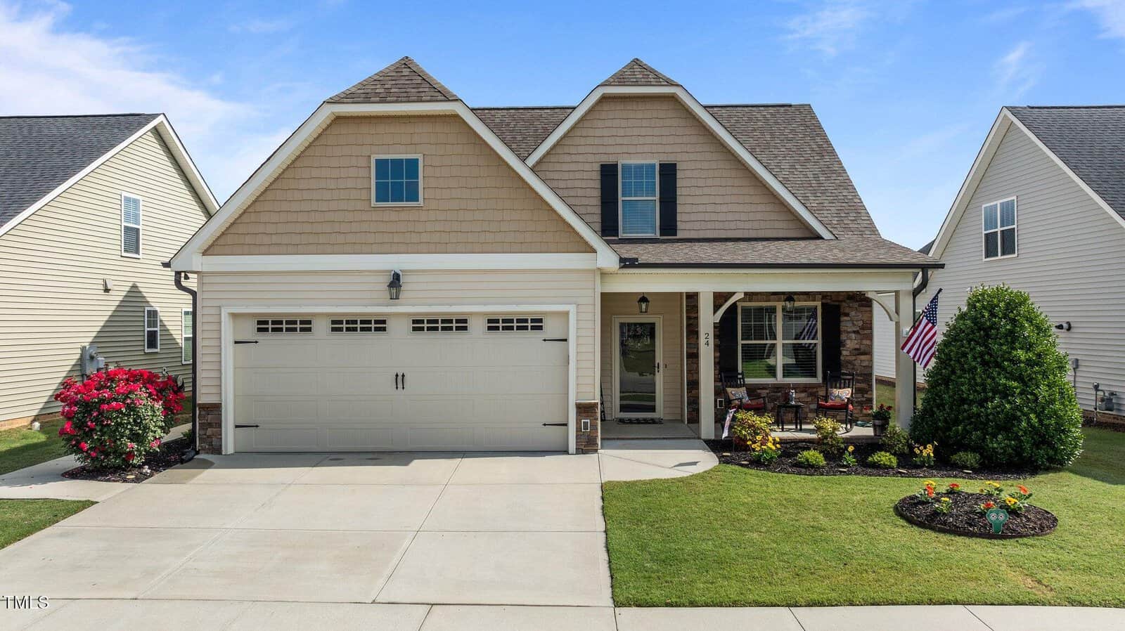 A beige suburban house with a double garage, gabled roof, front porch with chairs, and well-kept yard featuring flowering bushes and a small American flag.