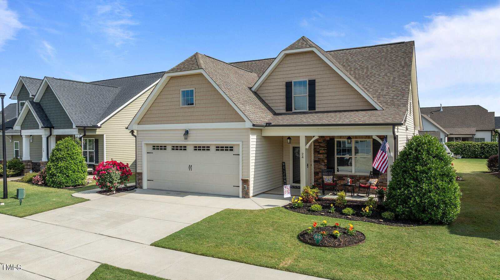 A suburban house with beige siding, double garage, front porch with chairs, a U.S. flag, and a well-kept lawn with flower beds, under a partly cloudy sky.