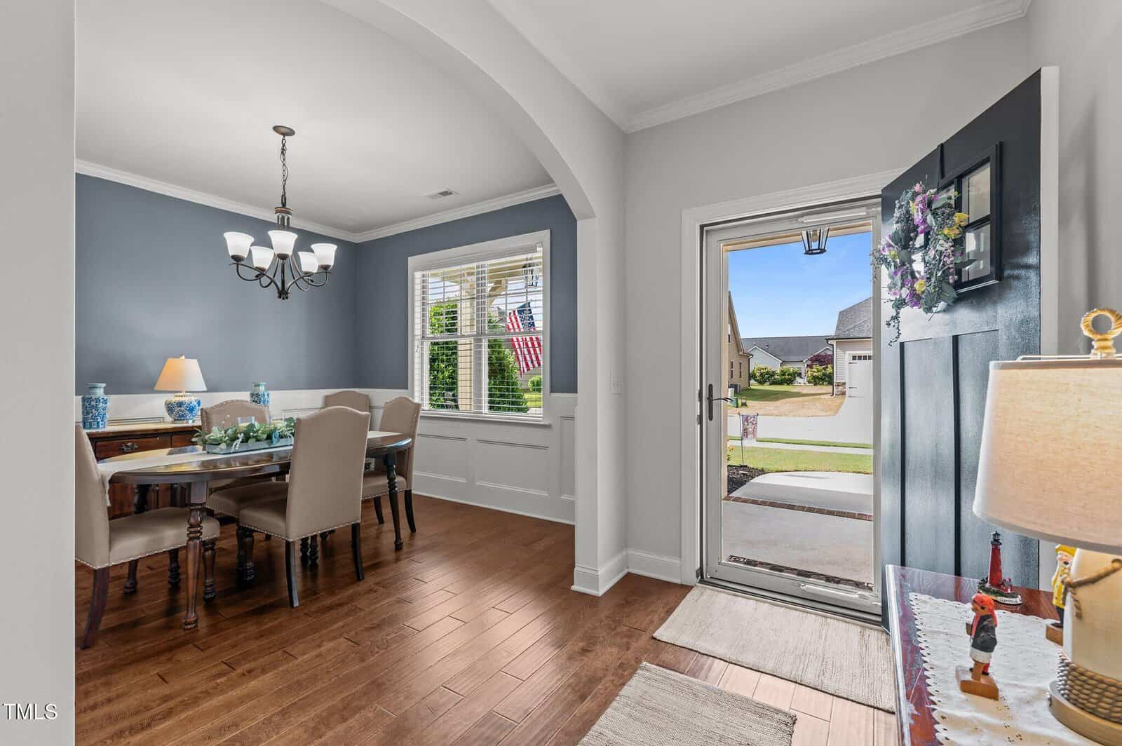 A front door opens to a bright entryway with a view of a dining room on the left, featuring wooden floors, a blue accent wall, beige chairs around a wooden table, and a window showing a U.S. flag outside.