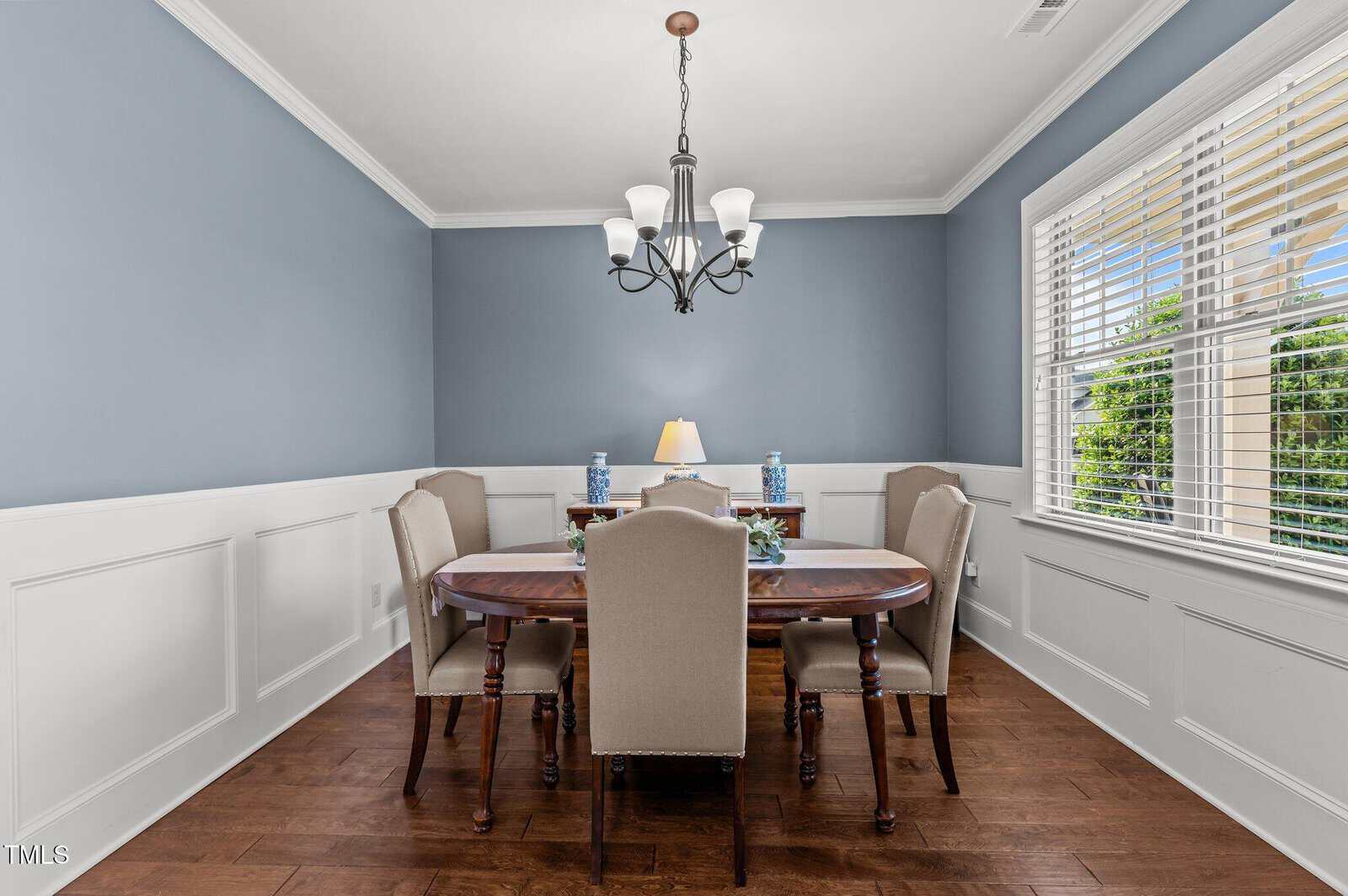 A dining room with blue walls, white wainscoting, a wooden floor, and a dark wood dining table surrounded by four upholstered chairs. A chandelier hangs above the table, and a window lets in natural light.