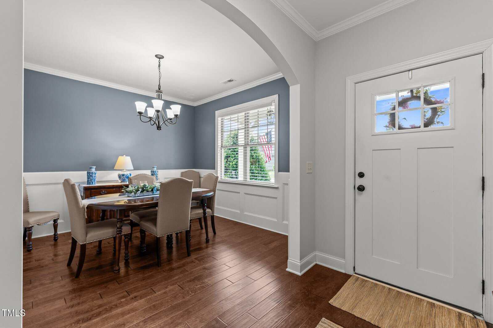 A bright dining room with blue accent wall, white wainscoting, wooden floor, a rectangular table with tan chairs, a chandelier, large window with blinds, and an entry door with glass window and decorative wreath.
