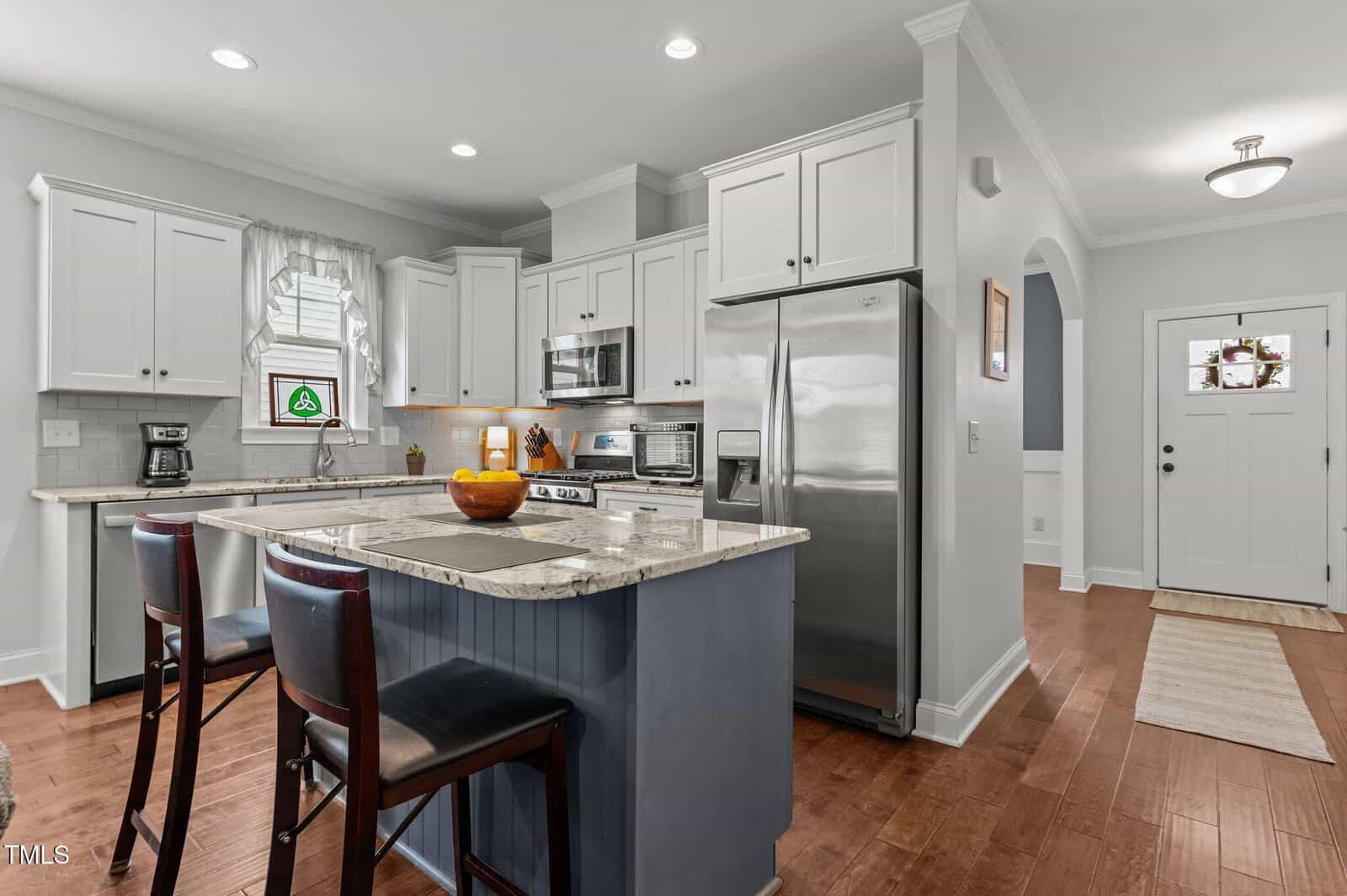 Modern kitchen with white cabinets, stainless steel appliances, and a center island with two chairs. Hardwood floors and a front entryway are visible, along with a bowl of oranges on the island.