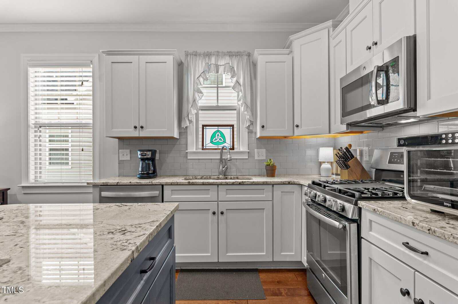 Bright kitchen with white cabinets, granite countertops, stainless steel appliances, a gas stove, a coffee maker, and a small window with a stained glass accent above the sink. Wood flooring and natural light are visible.