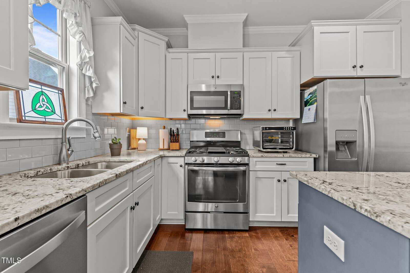 Modern kitchen with white cabinets, stainless steel appliances, a granite countertop, gas stove with microwave above, double sink under a window, and a refrigerator. A small potted plant and knife block are on the counter.
