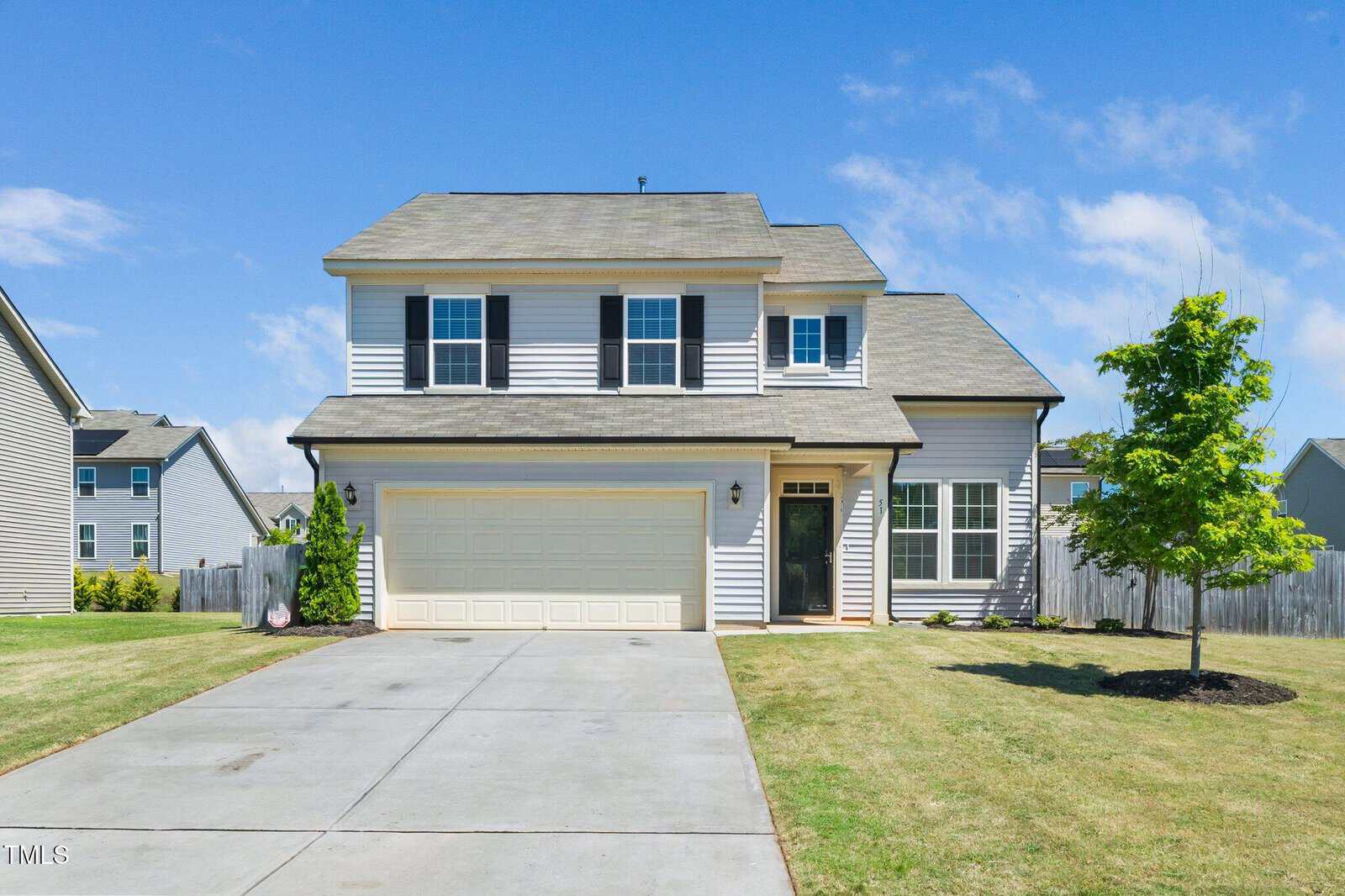 Two-story suburban house with light gray siding, dark shutters, a double garage, and a neatly manicured lawn. A small tree and some shrubs are planted near the front. The sky is blue with scattered clouds.