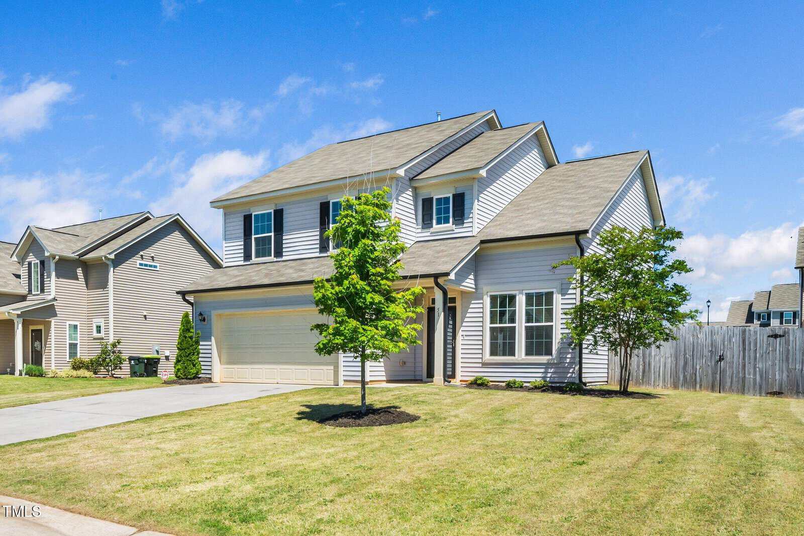 A two-story suburban house with beige siding, a two-car garage, and a well-maintained front yard featuring two small trees, under a blue sky with scattered clouds.