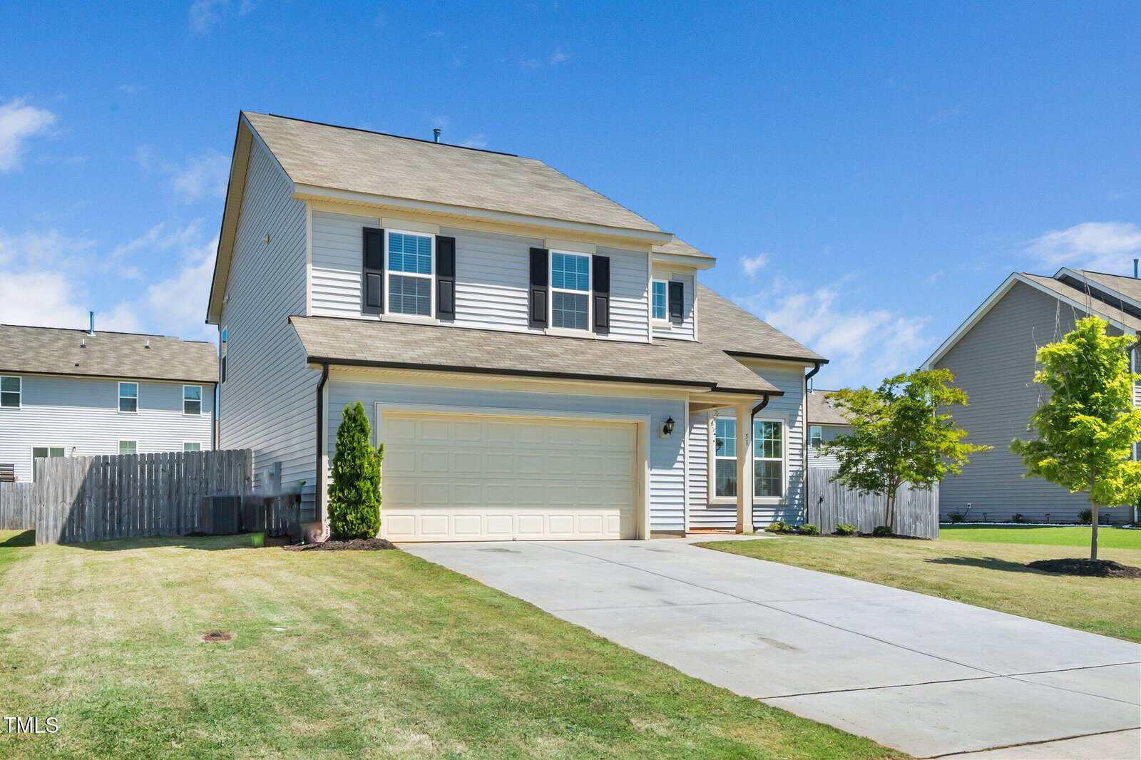 Two-story suburban house with beige siding, black shutters, and a two-car garage. The home has a large driveway, a green lawn, and is surrounded by a wooden fence and neighboring houses under a blue sky.