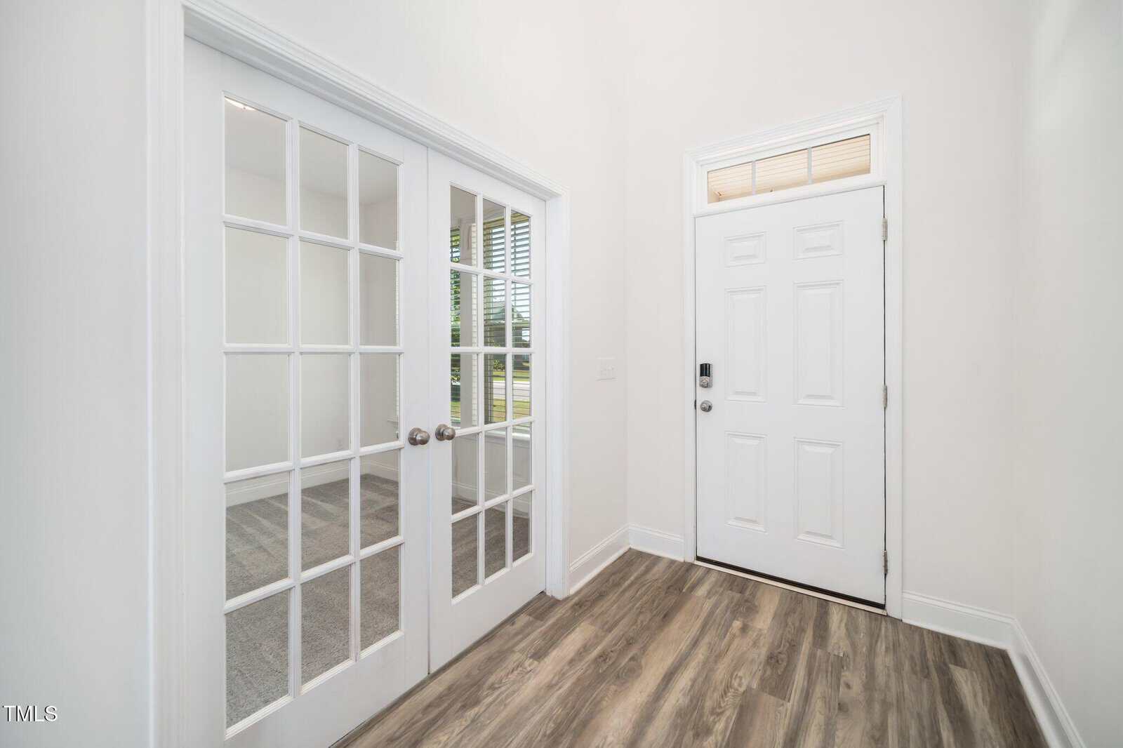 A bright, modern entryway with wood-look flooring, a white front door, and double French doors with glass panes leading to a room with carpet and windows.