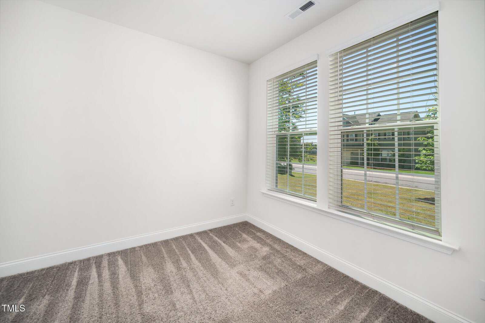 A bright, empty room with white walls, gray carpet, and two large windows with blinds, letting in natural light and offering a view of grass and a building outside.