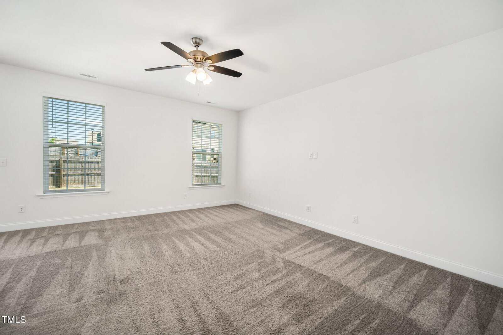 A clean, empty room with light gray carpet, two windows with blinds, white walls, a ceiling fan with lights, and visible vacuum lines on the carpet. Natural light enters through the windows.