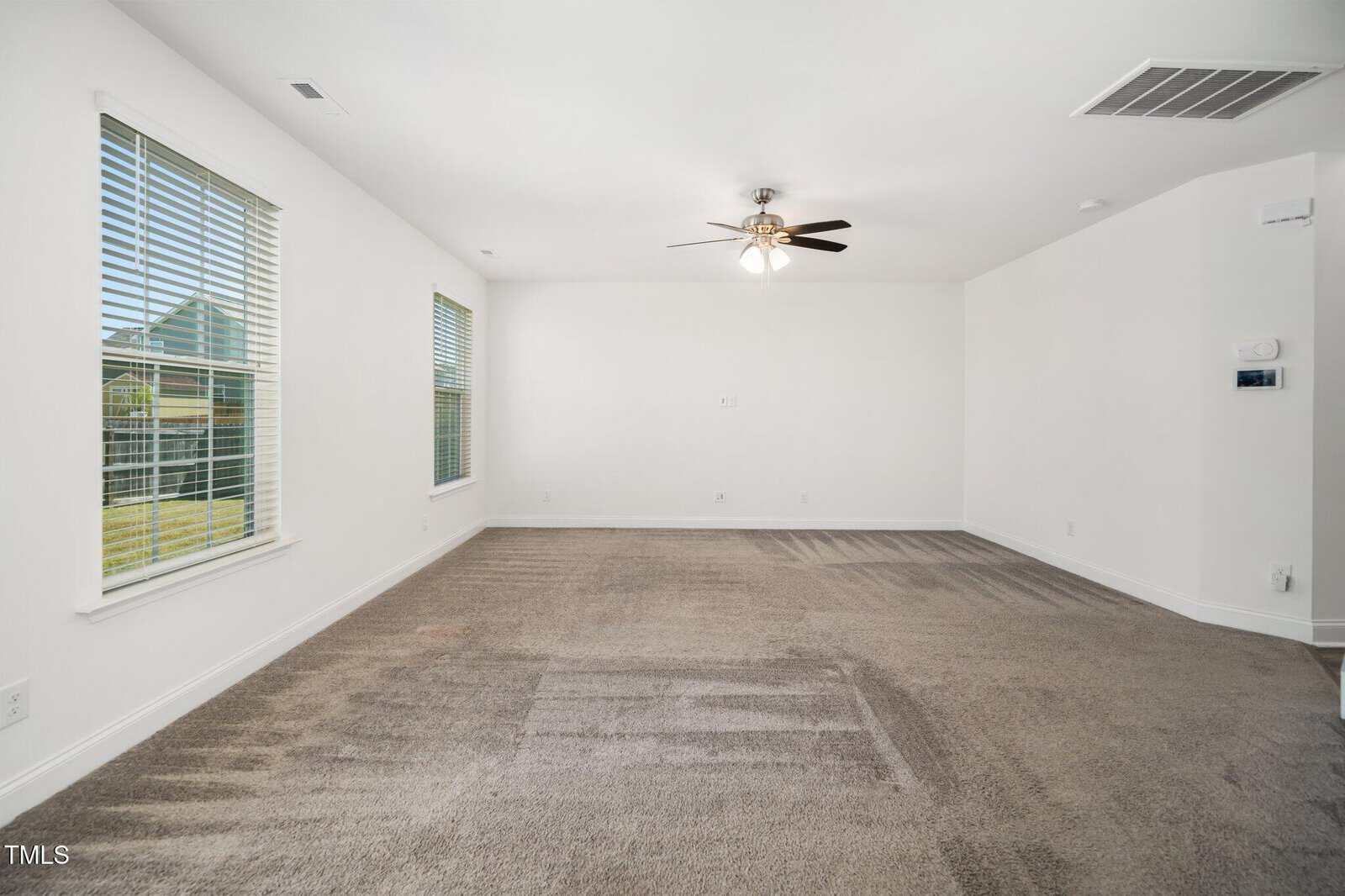 Empty, carpeted living room with white walls, three large windows on the left, a ceiling fan with lights in the center, and visible air vents and thermostat on the right wall. Sunlight is coming through the windows.