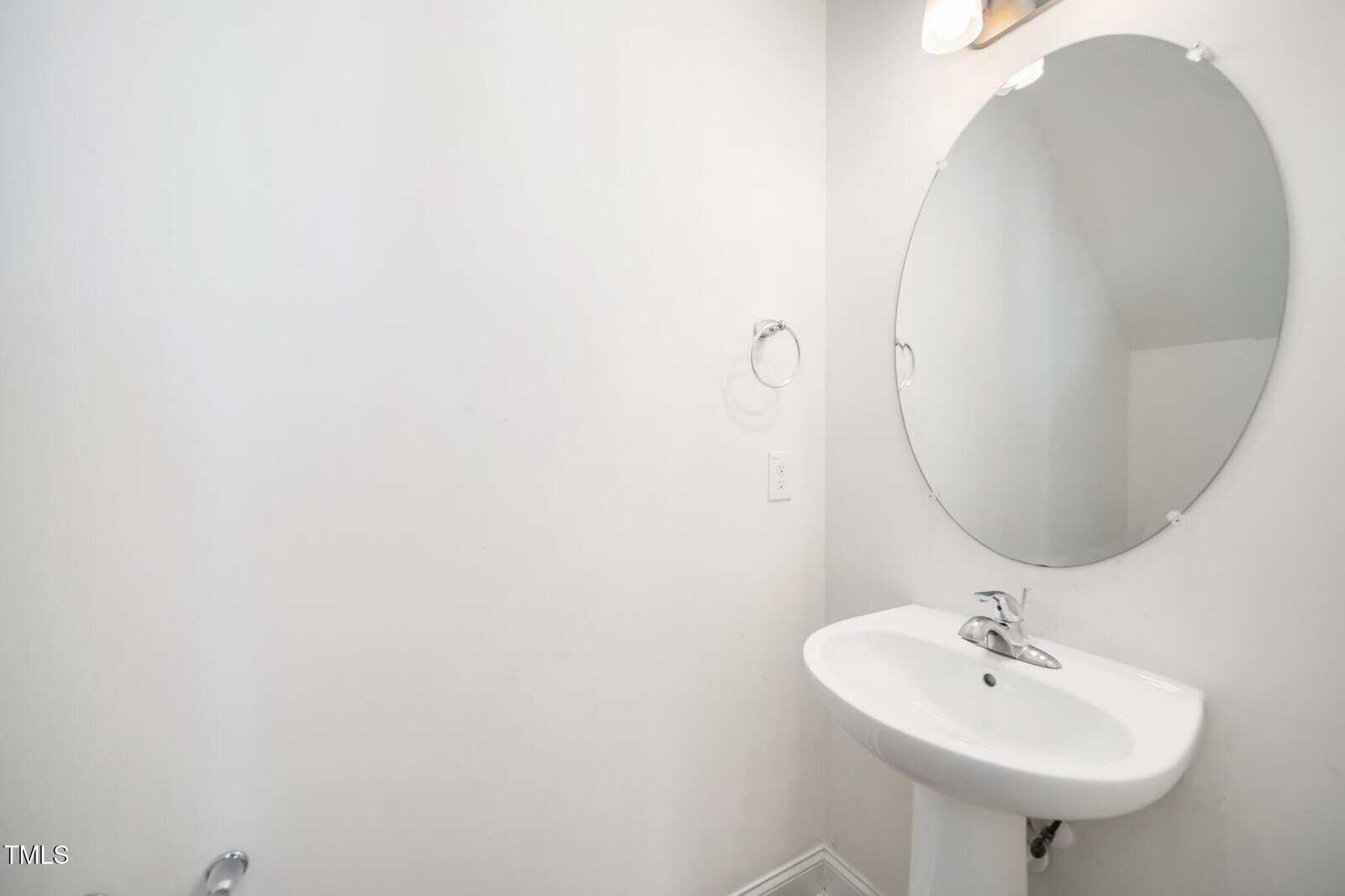 A small bathroom with white walls, an oval mirror above a white pedestal sink, a wall-mounted faucet, a light fixture above the mirror, and a metal towel ring on the wall.