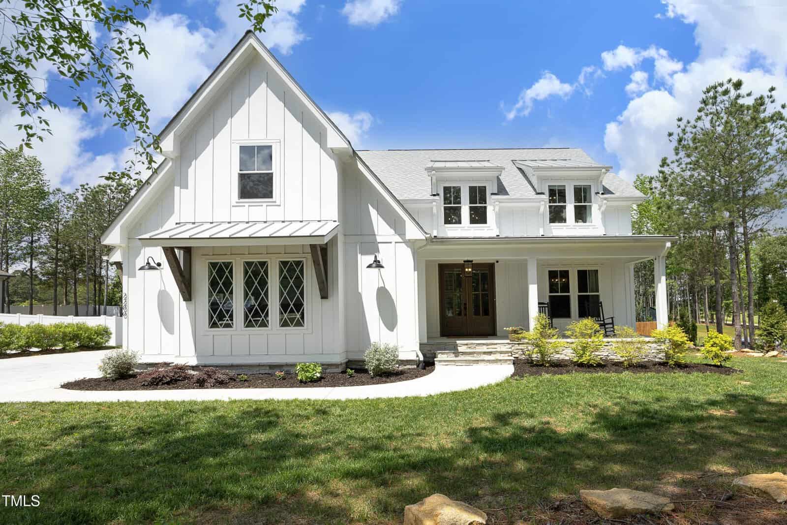 A modern white farmhouse-style home with large windows, a covered porch, and manicured landscaping, set against a backdrop of trees under a bright, partly cloudy sky.