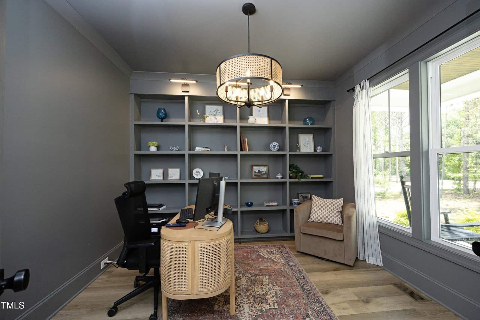 A modern home office with gray built-in shelves, a desk with dual monitors, a black office chair, a beige armchair, large windows with white curtains, and a patterned rug on light wood flooring.
