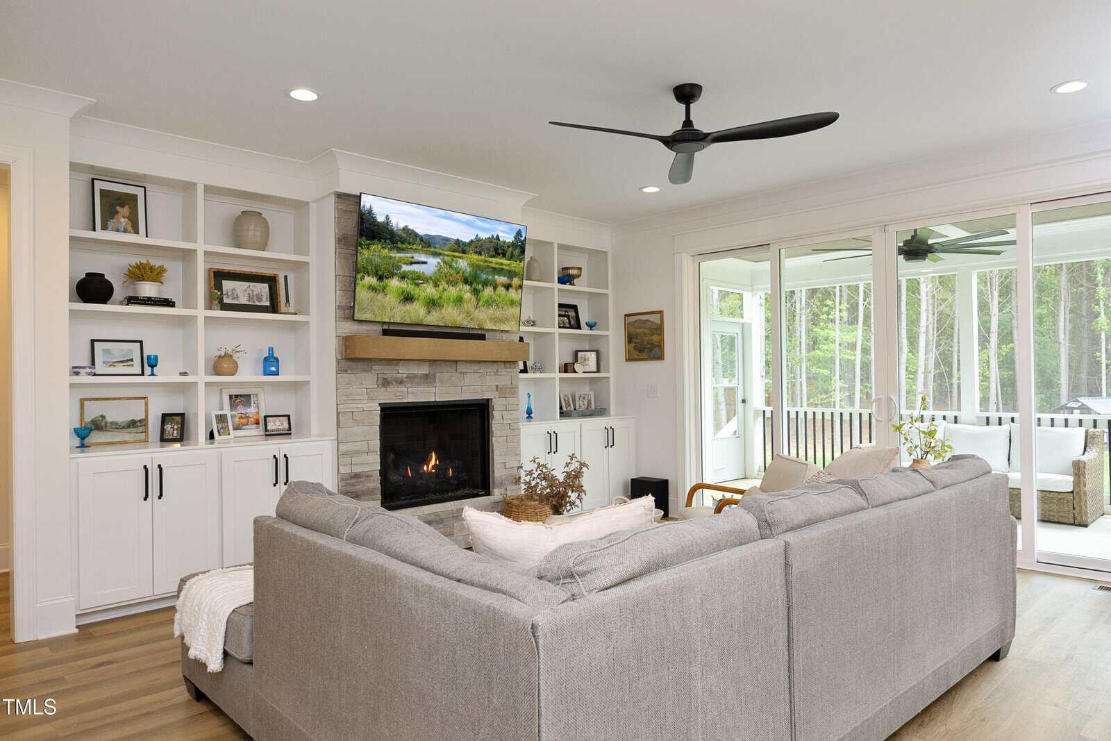 A modern living room with a gray sectional sofa, built-in shelves, a stone fireplace, a TV above the mantel, wood floors, a ceiling fan, and large windows leading to a sunroom with outdoor furniture.