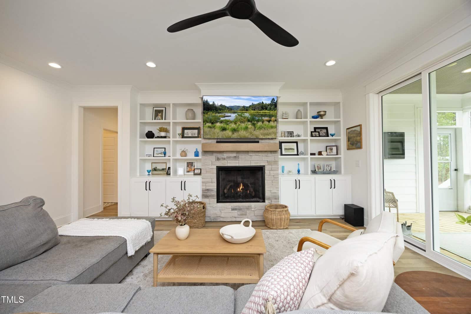 Modern living room with a gray sectional sofa, wooden coffee table, fireplace with stone surround, wall-mounted TV, built-in white shelves with decor, and sliding glass door leading to a patio. Ceiling fan overhead.