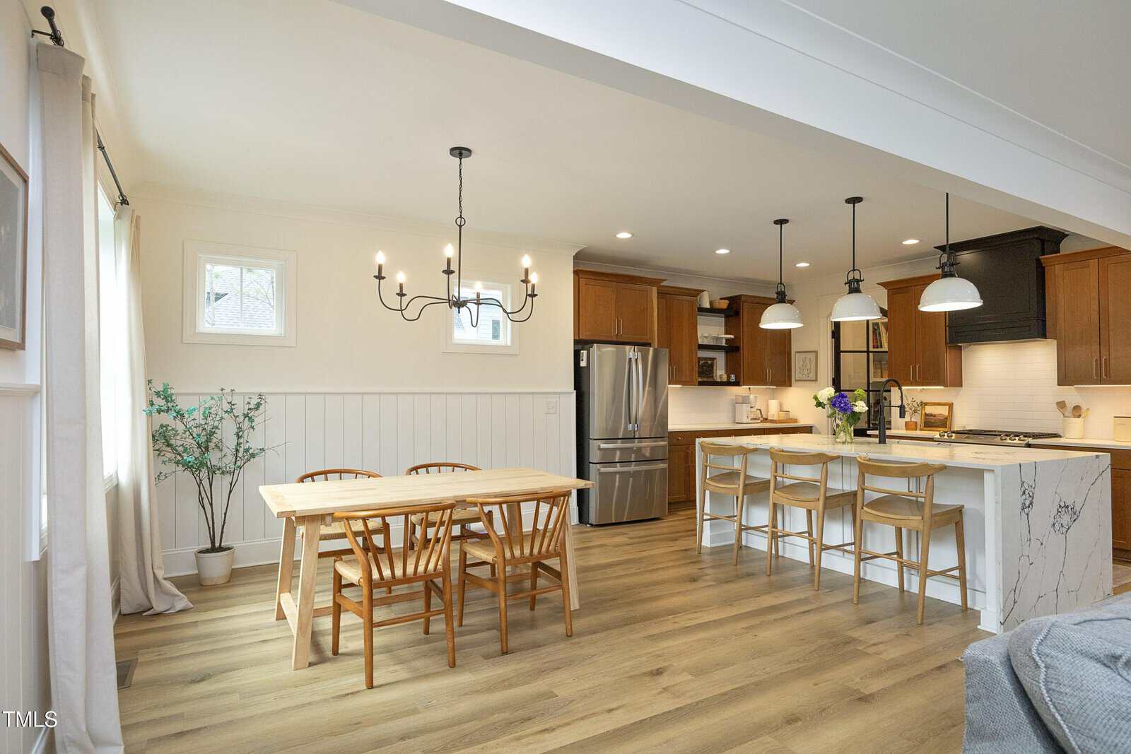 Bright kitchen and dining area with wood floors, wooden cabinets, a marble island with three stools, a wooden dining table with four chairs, pendant and chandelier lighting, potted plant, and natural light from windows.