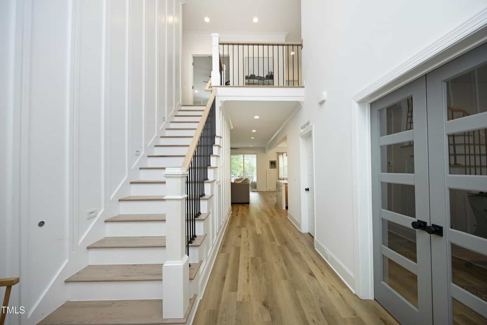A modern, bright foyer with light wood flooring, white walls, and a staircase with a black handrail leading to an open upstairs landing. Glass double doors are on the right, and the hallway leads to a kitchen area.