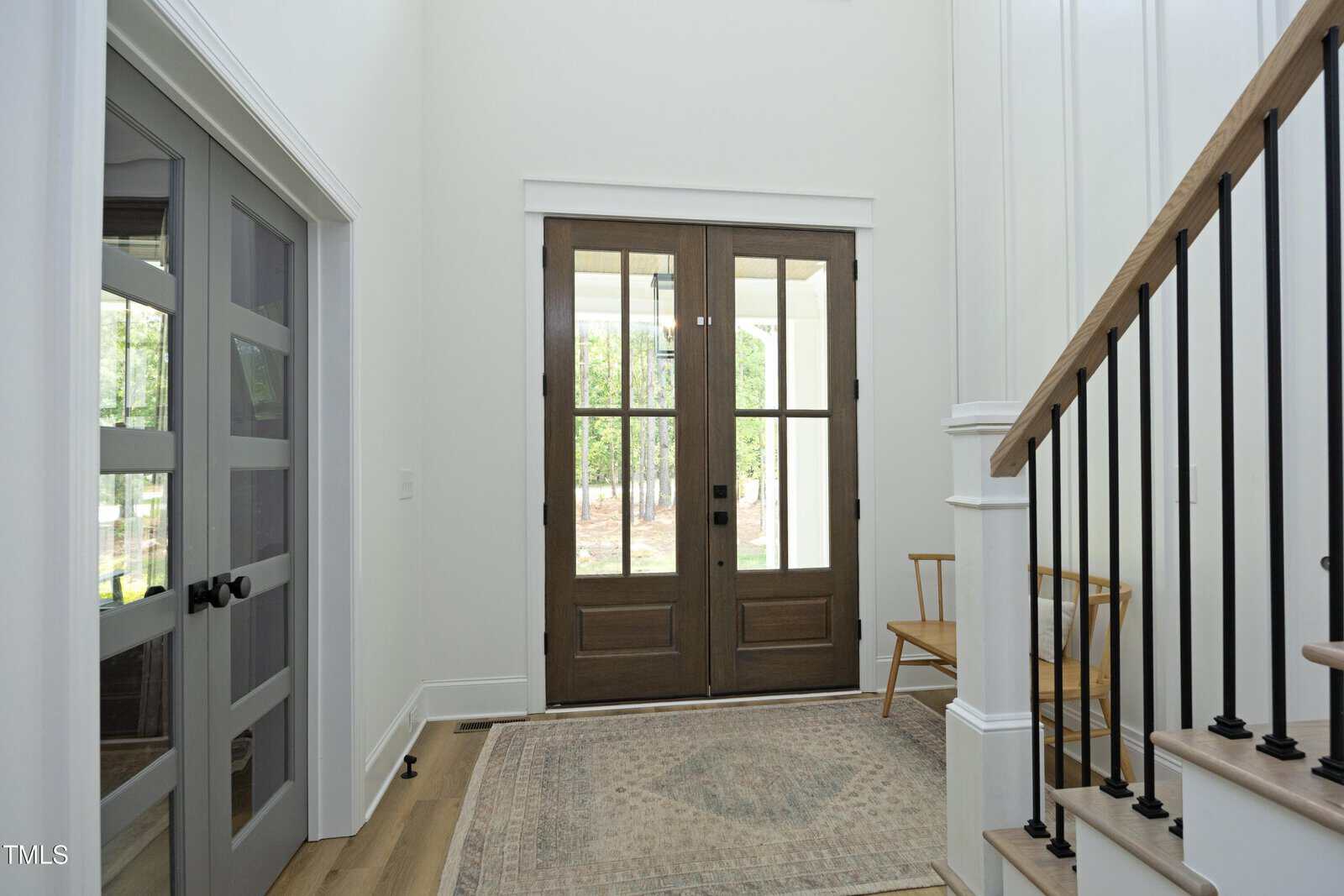 A bright entryway with double wooden front doors, glass panels, a light patterned rug, a wooden chair, and a staircase with black railings. Sunlight filters in, highlighting the minimalist decor.