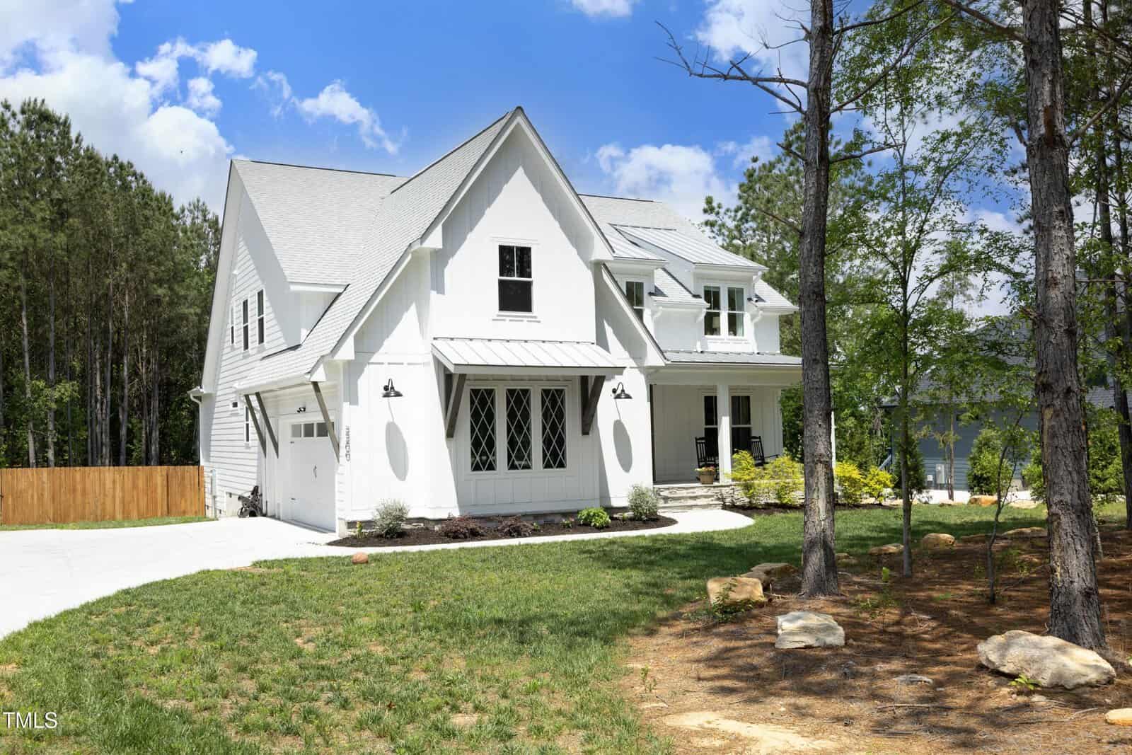 A modern white two-story house with large windows, a front porch, and a gray roof, sits on a spacious lawn with trees and a driveway, under a blue sky with scattered clouds.